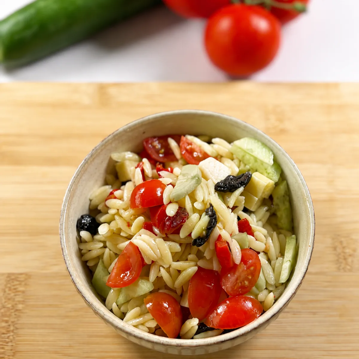 Schale mit Kritharaki-Nudelsalat, Cherrytomaten, Gurkenwürfeln, Gouda-Stückchen und schwarzen Oliven auf Holzbrett, im Hintergrund Gurke und Tomaten am Rispenzweig.
