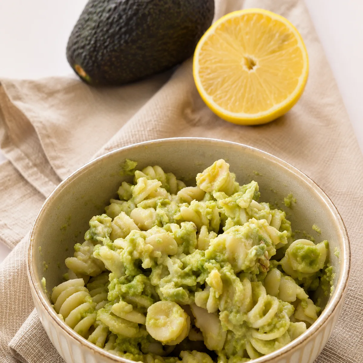 Schale mit Fusilli-Nudeln in cremiger grüner Avocado-Sauce auf Leinentuch, im Hintergrund eine ganze Avocado und eine halbierte Zitrone.